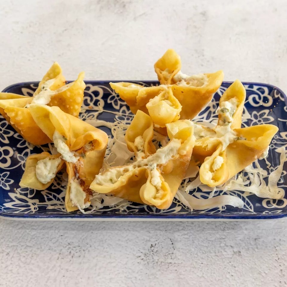 Crispy golden wontons filled with cream cheese and herbs, drizzled with white sauce on a blue plate.