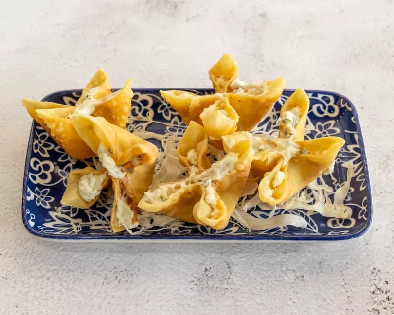 Crispy golden wontons filled with cream cheese and herbs, drizzled with white sauce on a blue plate.
