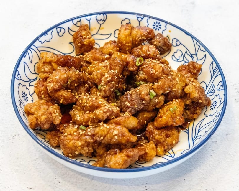 Crispy sesame chicken with green onions in a blue and white patterned bowl.