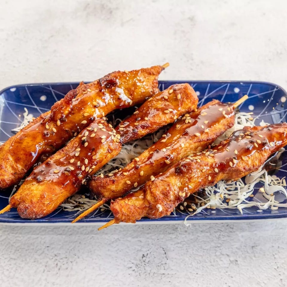 Six chicken skewers with teriyaki glaze and sesame seeds on a blue plate with shredded cabbage.