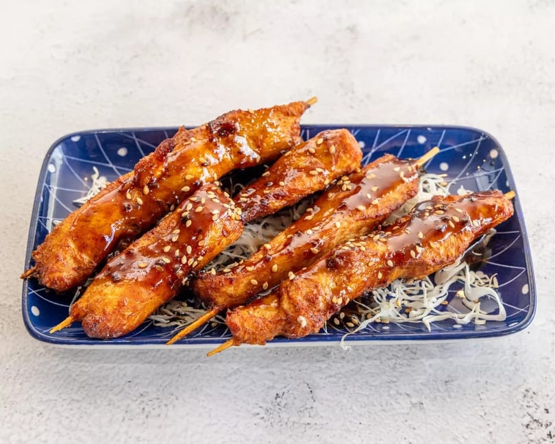 Six chicken skewers with teriyaki glaze and sesame seeds on a blue plate with shredded cabbage.