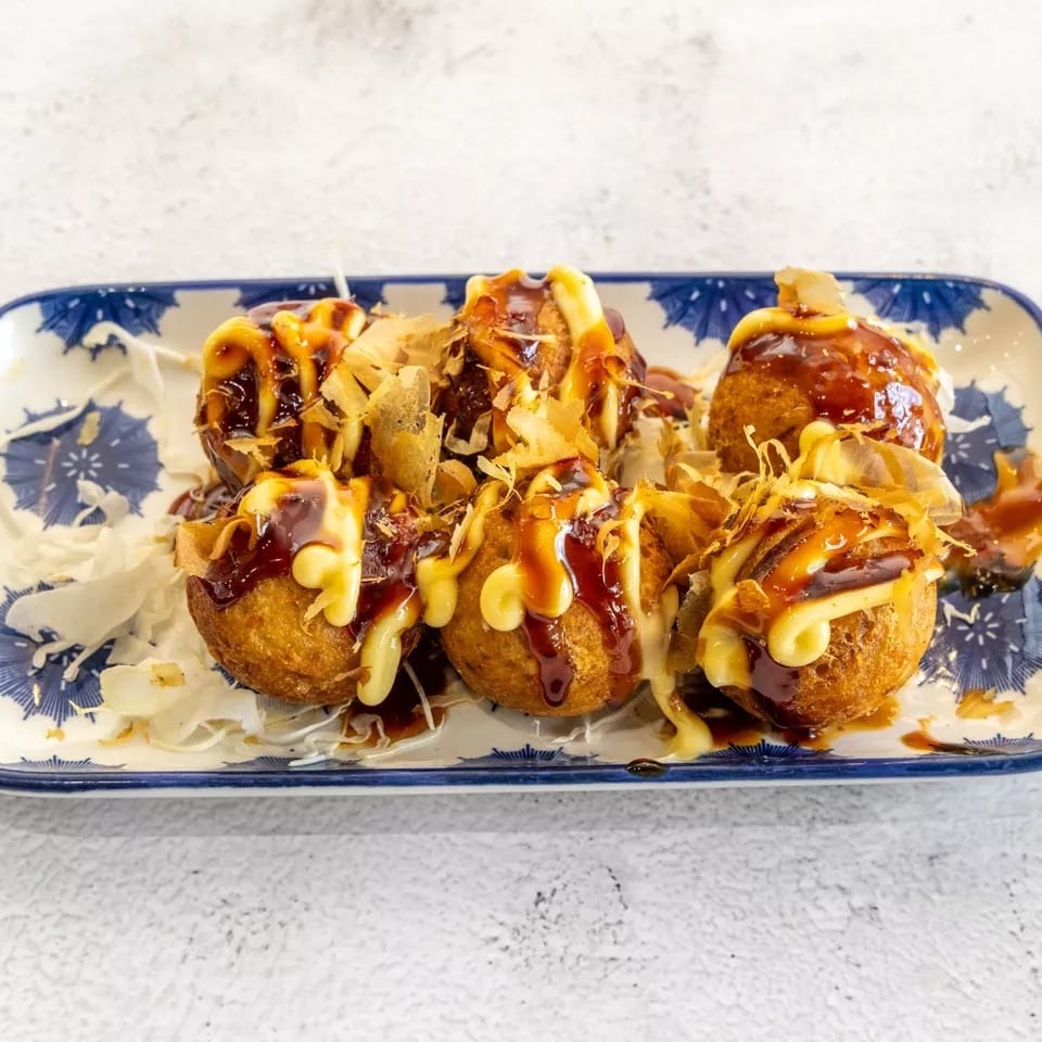 Six golden-brown takoyaki balls topped with dark sauce, mayonnaise, and bonito flakes on a blue and white plate.
