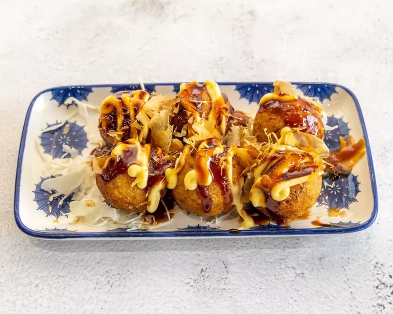 Six golden-brown takoyaki balls topped with dark sauce, mayonnaise, and bonito flakes on a blue and white plate.