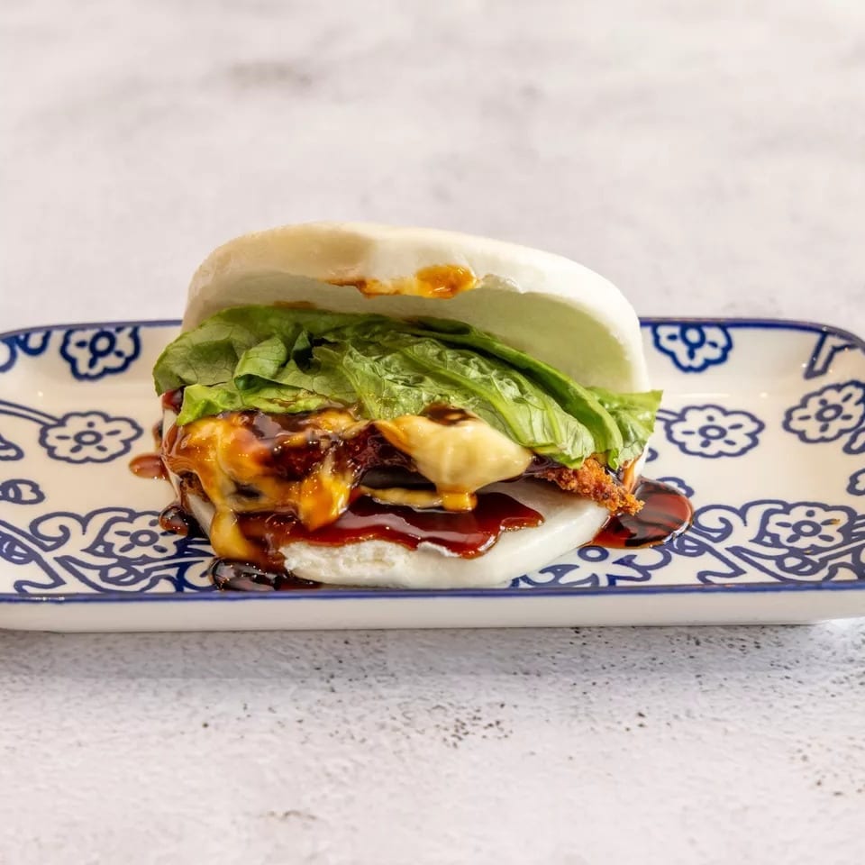 A bao bun filled with crispy chicken, melted cheese, lettuce, and dark sauce on a patterned plate.
