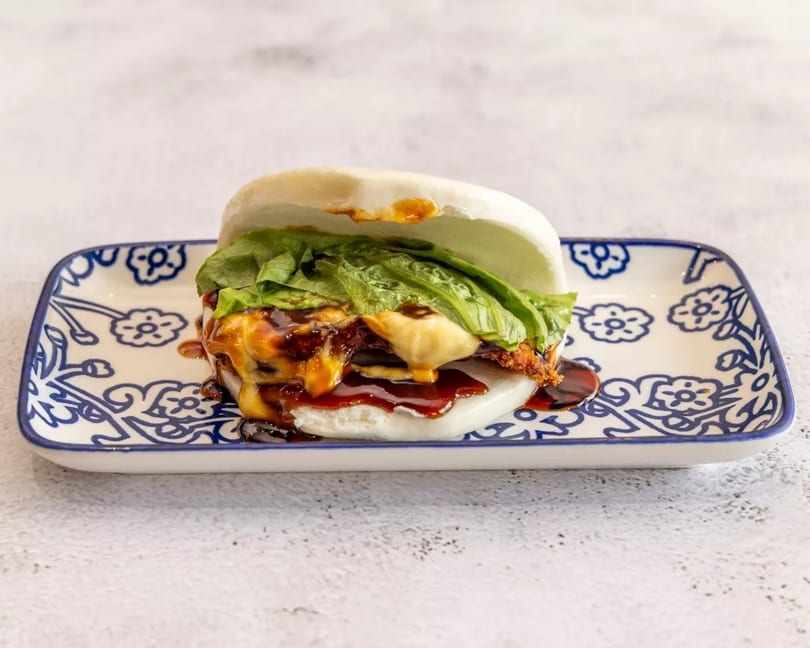 A bao bun filled with crispy chicken, melted cheese, lettuce, and dark sauce on a patterned plate.